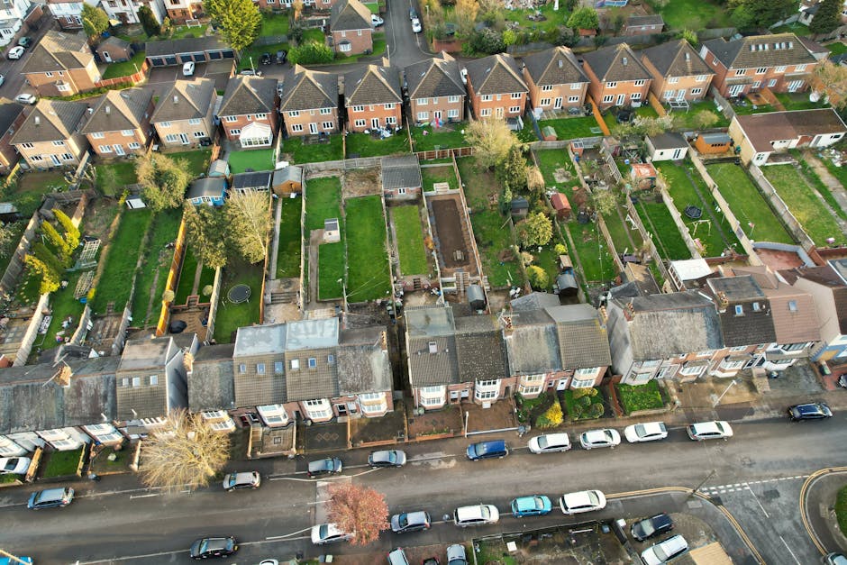 An aerial view of a suburban residential area showing a row of terraced houses with small front gardens and a street lined with parked cars in the foreground. Behind the houses, there are numerous pri
