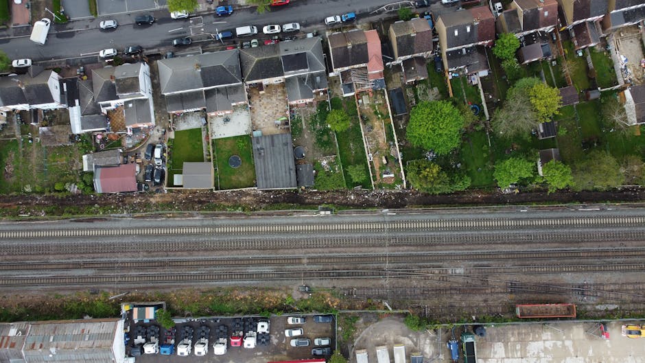 An aerial view of a row of residential houses with small gardens behind, featuring lush green trees, well-maintained lawns, and garden beds with various plants and shrubs. The gardens are bordered by 