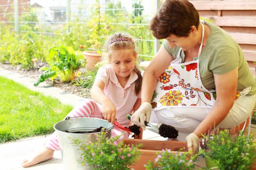 A woman with short dark hair, wearing a green top and floral apron, is kneeling on a paved outdoor area, engaging with her young daughter who has long hair tied back and is dressed in a light pink top and pink-striped trousers. They are both planting or tending to potted plants in rectangular garden planters, with gardening gloves on their hands. In the foreground, there are potted plants with green foliage and small purple flowers. Behind them, a lush garden with green grass, various plants, and a clear fence can be seen, indicating a well-maintained backyard in Catford. The scene is set under natural daylight, suggesting a pleasant, mild weather day, and highlights outdoor gardening activities typical of residential gardens in the area, supporting professional gardening services such as hedge trimming and landscape maintenance provided by Hedge Trimming Catford.